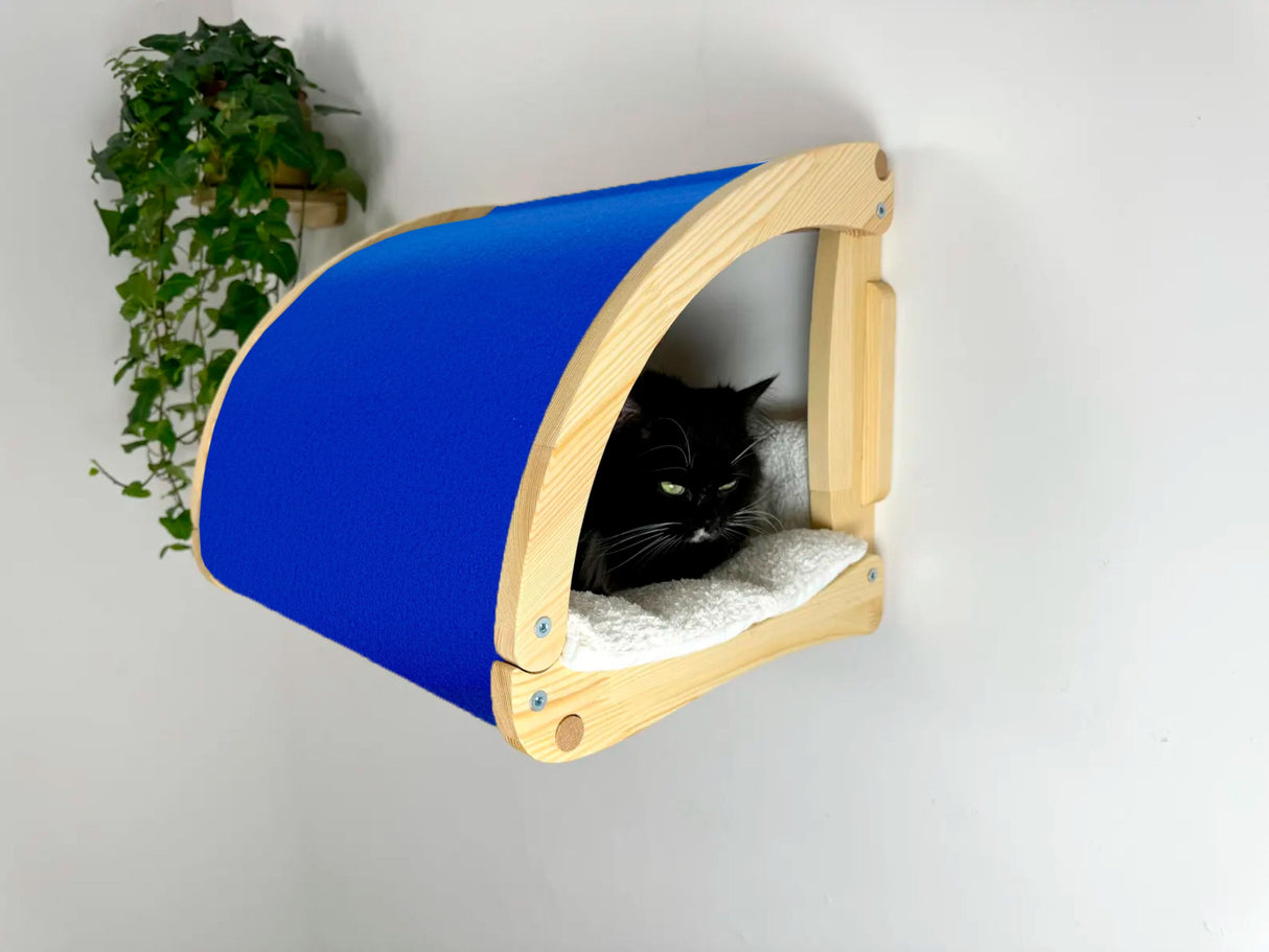 Blue and wooden cat bed with a black cat inside on a white wall.