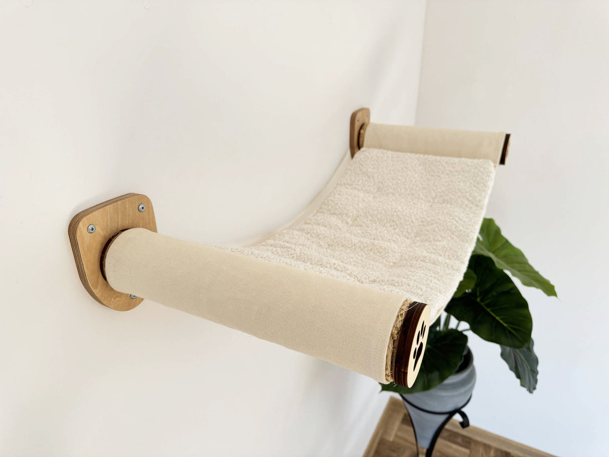 Wall-mounted cat hammock with wooden frame and beige cushion on a white wall.