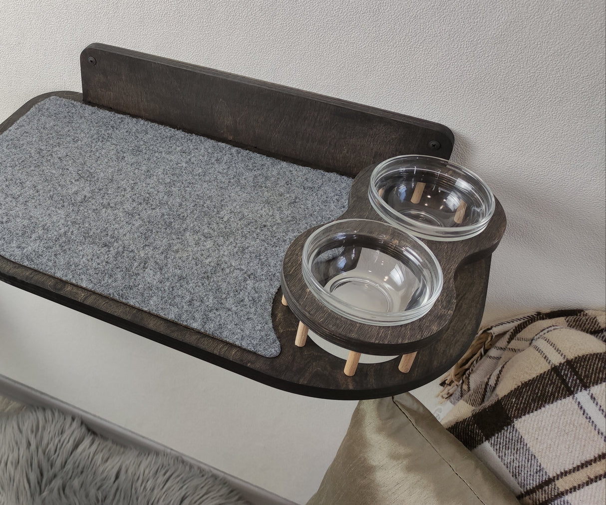 Wall-mounted cat feeder shelf crafted from dark wood, featuring a gray carpeted surface and two glass bowls for feeding