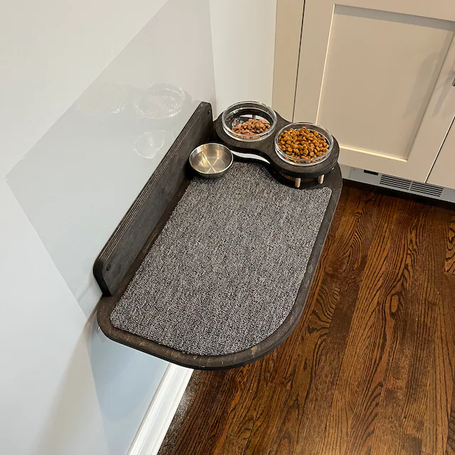 Practical and stylish cat feeder shelf made of dark wood, with a gray carpeted area and glass bowl set.