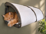Cat resting inside a cylindrical wooden and fabric cat bed attached to a wall.