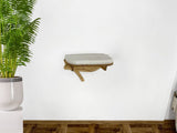 Wall-mounted cushioned cat bed against a white wall with a potted plant on the left.