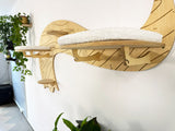 Wooden cat shelf with a cushion on a white wall, surrounded by plants.